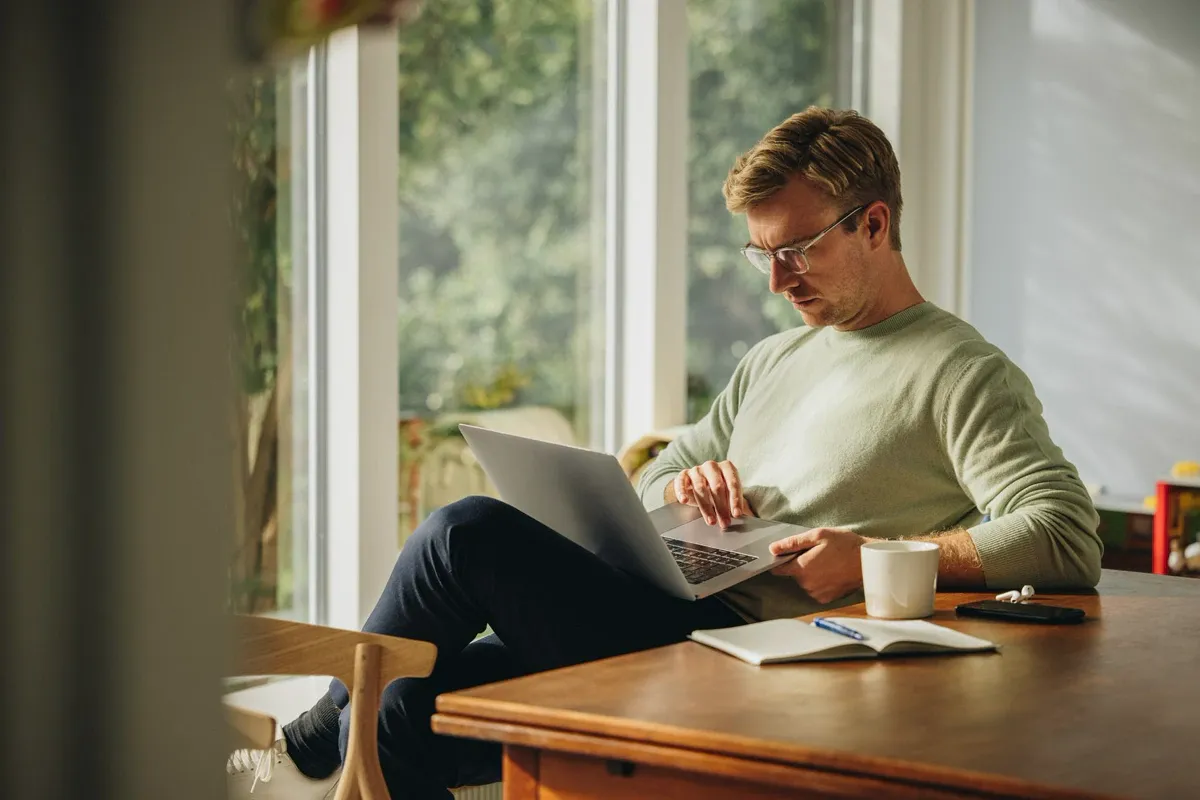 A man in glasses reading on a laptop beside a sunlit window, with a notebook and coffee on the table